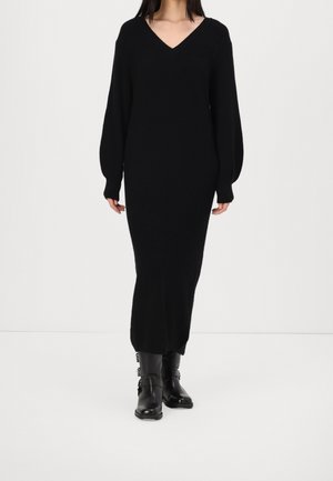 Femme portant une longue robe en maille noire à col en V avec des manches bouffantes et des bottes en cuir noires mi-mollet avec des boucles, debout sur un sol blanc.