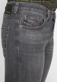Jeans de denim gris con un ligero estiramiento, que cuentan con cinco bolsillos y costuras en contraste. El bolsillo trasero muestra una etiqueta de marca.