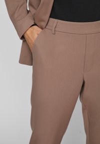 Pruunist suvekostüümist valmistatud püksid, mis on valmistatud siledast kangast, millel on lame esiosa, küljetaskud ja sobiv siluett. Kaasas bleiser.