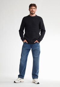 Zwarte gebreide trui met geribbelde manchetten, blauwe recht-toe-rechterschouder jeans, en witte sneakers met grijze accenten gedragen door een staande man.
