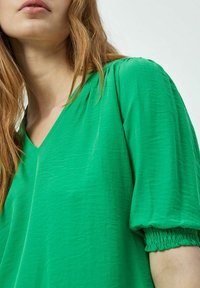 Blusa verde a scollo a V con maniche corte arricciate. Tessuto morbido e texture. Finitura liscia, design leggero. Dettaglio in primo piano del collo e delle maniche.