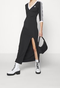 Svart maxiklänning med V-ringning och trekvartsärmar, med vita ränder på ärmarna och en hög slits på låret. Matchad med vita snörstövlar.