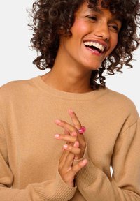 Beige Strickpullover mit langen Ärmeln, der über einen gerippten Ausschnitt und Bündchen verfügt. Das Modell trägt einen pinken Ring und hat die Hände vor sich gefaltet.