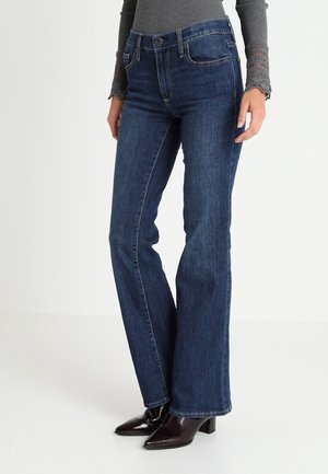 Frau trägt dunkelblaue Bootcut-Jeans, graues Langarmshirt mit Spitzenmanschetten und schwarze, hochhackige Stiefeletten, steht vor einfachem Hintergrund.