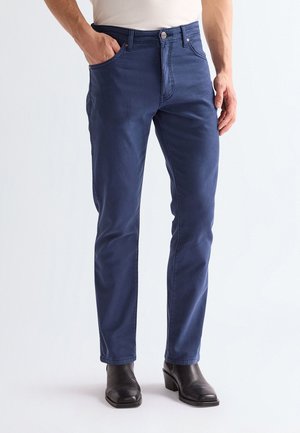 Mand iført marineblå slim-fit bukser og sorte læderankelstøvler, med hånden i lommen, der står mod en ensartet lys baggrund.