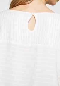 Bílá blůza s texturovaným vzorem a vertikálními pruhy, kulatým výstřihem a klíčovou dírkou na zádech uzavřenou knoflíkem.