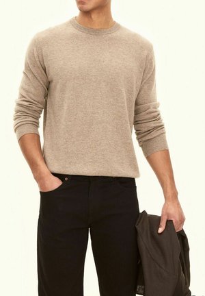 Maglione beige a maglia con collo rotondo e polsini a coste, abbinato a pantaloni neri. Il modello tiene una giacca scura in una mano.