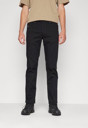 Homme portant un pantalon noir slim, des baskets noires et une chemise beige à manches courtes sur un fond clair uni.