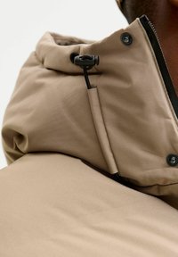 Lähikuva beigestä takin kauluksesta, jossa on musta kiristysnauhan säätö ja nepparit, jota kantaa tummaihoinen henkilö.