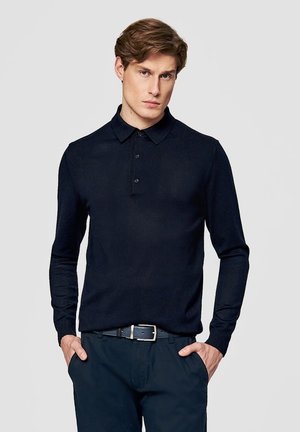 Młody mężczyzna o jasnobrązowych włosach, ubrany w czarną koszulkę polo z długim rękawem i ciemne spodnie, z rękami w kieszeniach, na jednolitym tle.