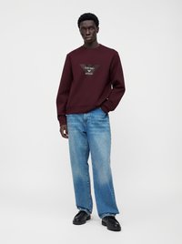 Мъж, облечен с бордо суитшърт на Emporio Armani, сини дънки и черни обувки, стои на бял фон.