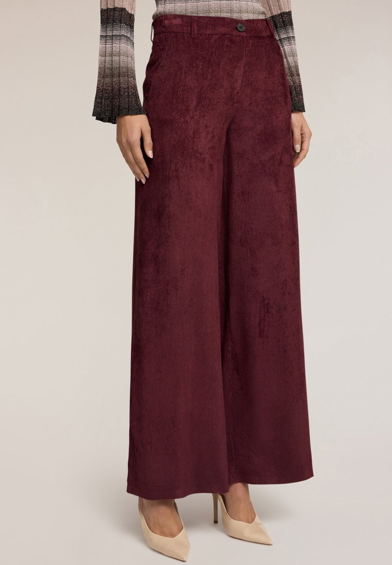 Pantalones de pierna ancha en burdeos, fabricados en un tejido suave con una textura lisa, que cuentan con un cierre delantero de botón y bolsillos laterales. Combinados con tacones nude.