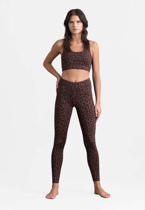 Frau steht barfuß, trägt einen braunen Sport-BH und Leggings mit Leopardenmuster, Hände in der Hüfte, vor einem weißen Hintergrund.