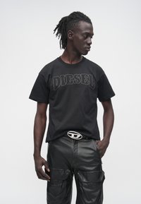 Čierne bavlnené tričko s nápisom "DIESEL" v kontrastnom prešívaní, doplnené čiernymi koženými cargo nohavicami s viacerými vreckami a lesklou prackou opaska.