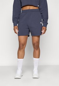 Calções de algodão azul-marinho com um cós elástico, bolsos laterais e um pequeno logótipo na bainha. Combinados com meias brancas e ténis.