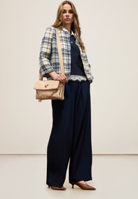 Geverfde blauwe en witte tweedjas over een marineblauw topje, gecombineerd met brede marineblauwe broek en een beige handtas met gouden kettingdetail. Bruine hakken.