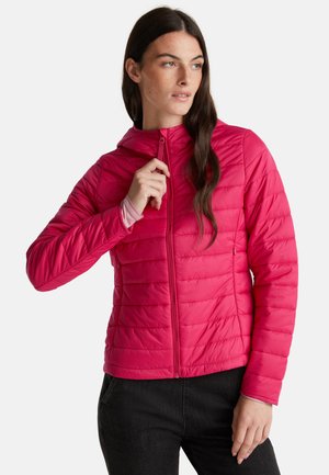 Vrouw die de rits van een felroze gewatteerde jas met capuchon aanpast, gekleed in een zwarte broek en een lichtroze langslavende shirt eronder.