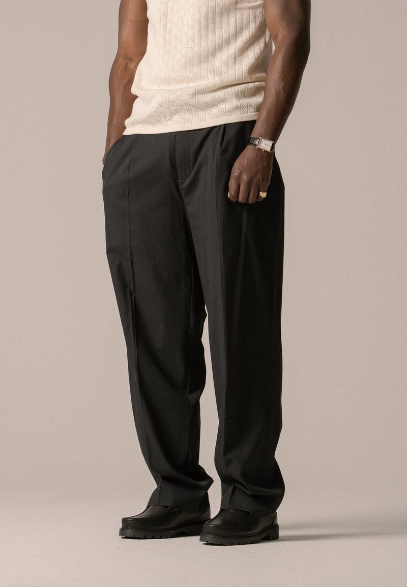 Pantalon noir à jambes larges avec une texture lisse, doté de plis à l'avant, porté avec des chaussures noires. Le tissu semble léger et fluide.