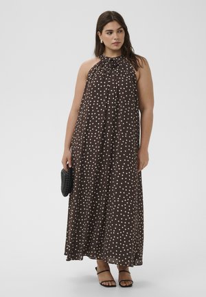 Žena v hnedých bezrukávových maxi šatách s bodkami, držiaca čiernu kabelku, obutá v čiernych sandáloch a s perlovými náušnicami, stojaca pred jednofarebným pozadím.