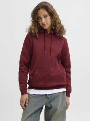 Burgunderfarget hettegenser med kengurulomme, justerbar snorhette og ribbestrikkede mansjetter, lagd over en hvit skjorte, kombinert med løse jeans.