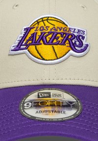 Regulējams pelēkas un purpursarkanas krāsas cepures modelis ar izšūtu "Los Angeles Lakers" logo un basketbola dizainu priekšpusē; redzama New Era zīmola birka.