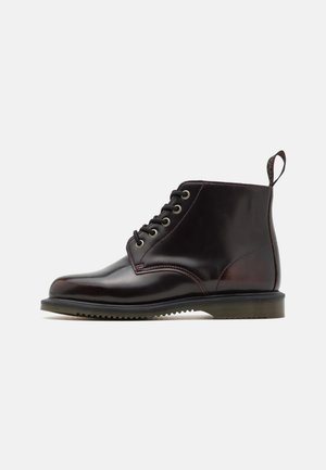 Botas de tornozelo em pele castanha com bico redondo, design com atacadores, ilhoses metálicos e sola texturizada. Apresenta uma alça na parte de trás.