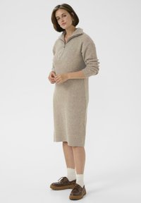Een vrouw staat in een beige ribgebreide midi-jurk met een kwartrits-kraag, witte sokken en een paar bruine veterschoenen op een effen achtergrond.