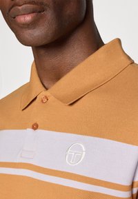 Pomarańczowa koszulka polo w białe poziome paski, z teksturowanej tkaniny, z zapięciem na trzy guziki i okrągłym logo na piersi.
