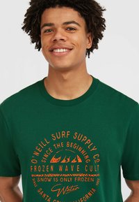 Grøn bomulds t-shirt med orange grafisk tekst og bølgemønster på forsiden. Klassisk rund hals og korte ærmer.