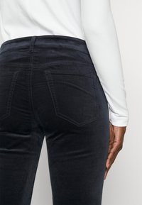 Personne portant un pantalon noir ajusté et un haut blanc à manches longues, vue de dos de la taille jusqu'au milieu de la cuisse, la main reposant le long du corps.