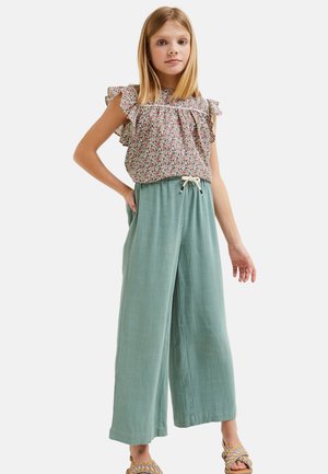 Jeune fille portant un haut à manches courtes volantées à motif floral, un pantalon vert ample avec cordon de serrage, et des sandales tressées, debout avec une main sur la hanche.