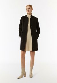 Schwarzer Mantel mit geradem Schnitt, zwei Vordertaschen und Knopfverschluss, getragen über einem beigefarbenen Kleid und hellen Knöchelstiefeln.