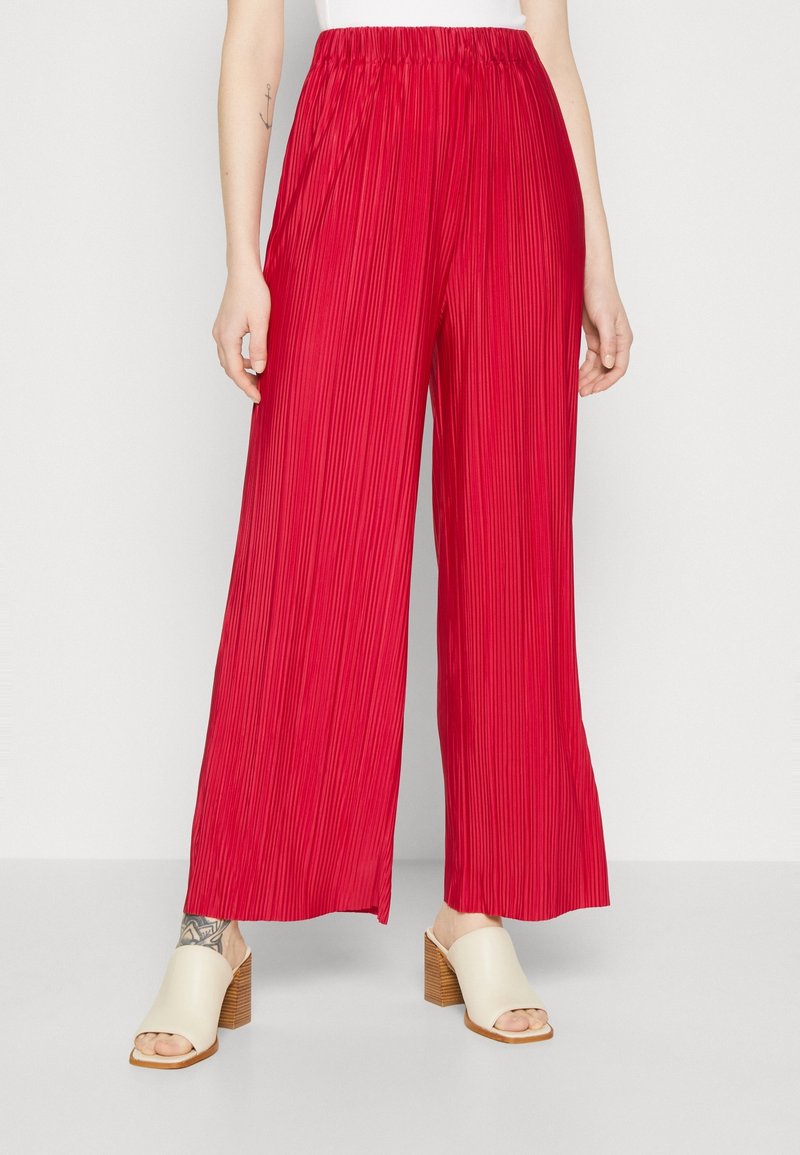 Femme portant un pantalon large plissé rouge vif et des sandales crème à talons blocs ouvertes à l'avant, debout devant un fond uni.