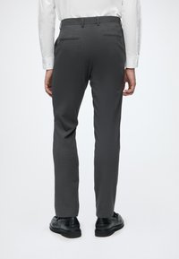 Homme portant un pantalon formel gris foncé, des chaussures habillées noires et une chemise blanche à manches longues, debout de dos sur un fond blanc.