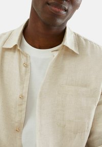 Beige linnen overhemd met een kraag, knoopsluiting en een borstzak. Eronder is een witte t-shirt zichtbaar. Zachte, gestructureerde stof.
