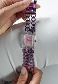 Montre carrée Guess avec un boîtier transparent, un cadran rose et un bracelet en maillons métalliques multicolores. Le bracelet présente un design audacieux et structuré.