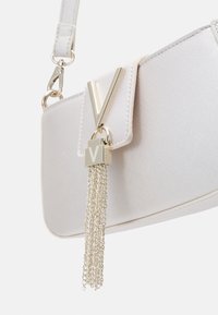 Bílá syntetická kabelka s texturovaným povrchem, vybavená zlatým V-tvarovaným uzávěrem a stříbrným detailním střapcem. Součástí je nastavitelné poutko.