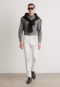 Siva košulja s dugim rukavima, bijele slim-fit hlače, crni džemper prebačen preko ramena, crne sunčane naočale, crne cipele, minimalistička pozadina.