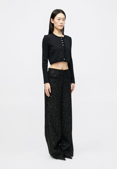 Femme debout portant un cardigan noir à boutons et à manches courtes, associé à un pantalon pailleté évasé taille haute et des chaussures noires à bout pointu.