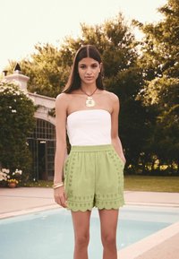 Helle grüne, gewellte Shorts mit bestickten Details und Seitentaschen, kombiniert mit einem weißen trägerlosen Oberteil. Poolside-Umgebung.