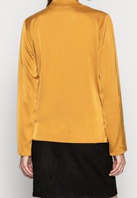 Żółta bluzka z satyny z długim rękawem, kołnierzykiem oraz gładką, gładką fakturą. Stylizowana z czarną spódnicą o prostym kroju.