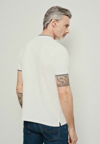 Biała koszulka polo z krótkim rękawem, z czarnymi paskami na kołnierzyku i mankietach. Z bocznymi rozcięciami dla wygody. Na ramieniu widać tatuaże.