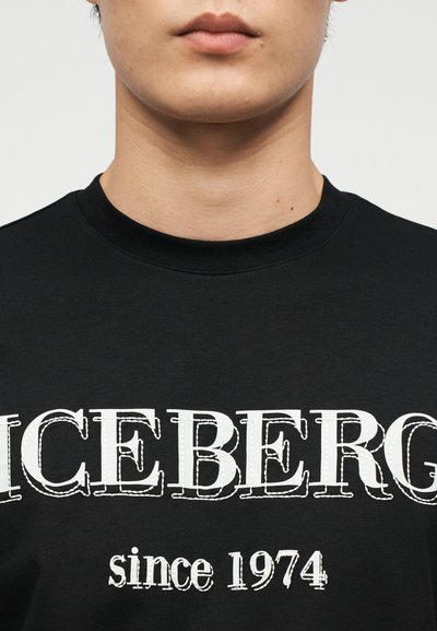 Κοντινό πλάνο του κάτω μέρους του προσώπου και του λαιμού ενός ατόμου που φοράει μαύρο μπλουζάκι με στρογγυλή λαιμόκοψη και το λευκό κείμενο "ICEBERG since 1974".