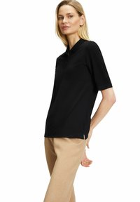 Zwarte polo met korte mouwen en een kraag, voorzien van een knoopsluiting aan de voorkant. Het shirt heeft een ontspannen pasvorm en zijsplitten voor meer bewegingsvrijheid.