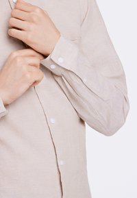 Camicia beige con bottoni, realizzata in tessuto testurizzato, con bottoni bianchi e una singola tasca sul petto. Il polsino è leggermente arrotolato.