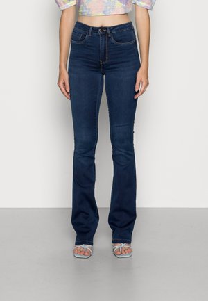 Kvinde iført mørkeblå jeans med høj talje og bootcut samt lyseblå sandaler med stropper, der står foran en ensfarvet baggrund.