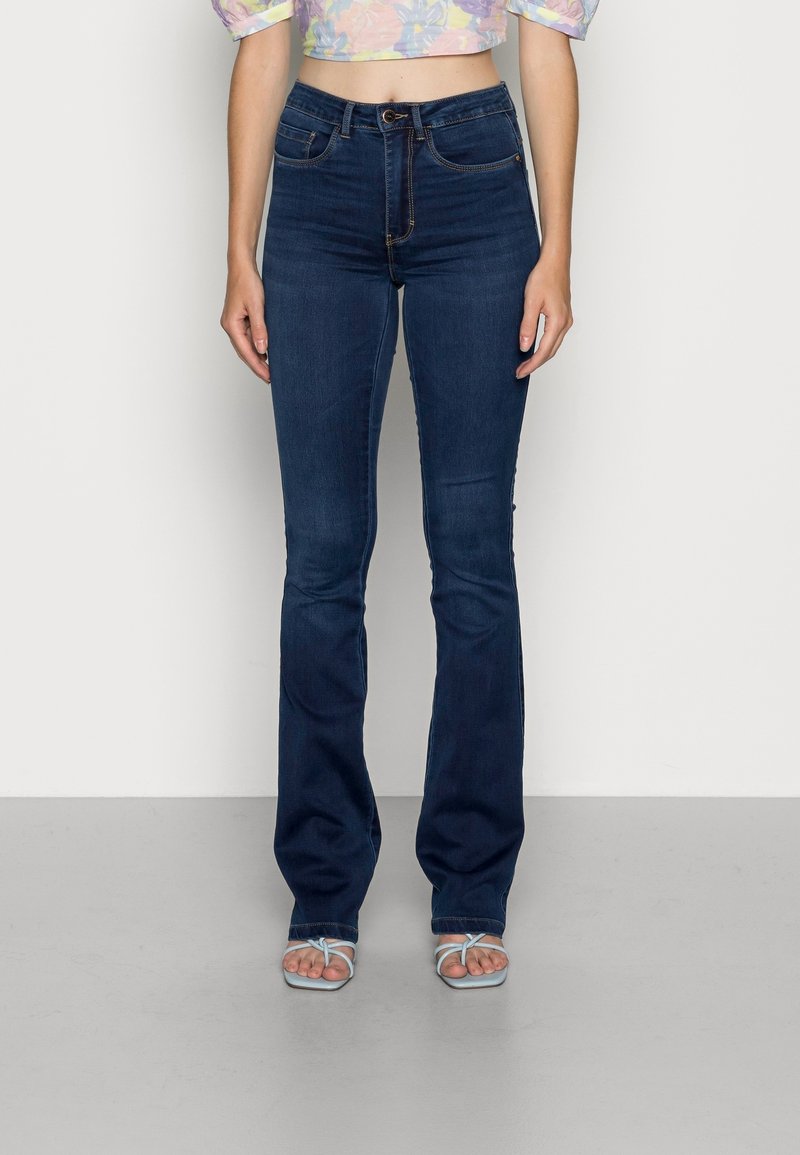 Femme portant un jean bootcut taille haute bleu foncé et des sandales à lanières bleu clair, debout devant un fond uni.