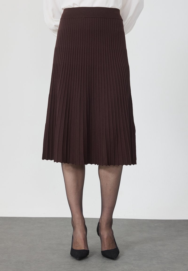 Personne portant une jupe midi plissée marron foncé, des collants noirs transparents et des escarpins noirs à bout pointu, debout sur un sol gris.