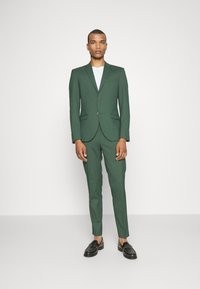 Zeleni elegantni kostim s slim fit krojem, dvema gumboma, notch ovratnikom in enim žepom na prsih, kombiniran z črnimi loaferji in belo srajco.