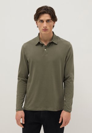 Giovane uomo con capelli castani mossi, indossa una polo verde oliva a maniche lunghe e pantaloni neri, in piedi davanti a uno sfondo uniforme.